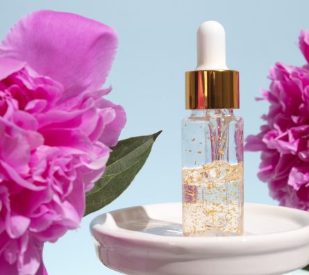 Glass dropper bottle on podium with peony flowers on blue background. Cosmetic container mock-up. Glass dropper bottle on podium with peony flowers on blue background. Cosmetic container mock-up.